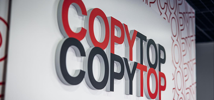 Logo COPYTOP composé de grandes lettres rouges, grises et noires en relief, fixé sur un mur blanc dans un espace professionnel moderne.