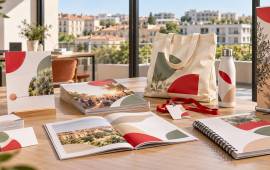 Brochures, cartes, dossier de présentation, badge, tote bag et gourde dans un bureau élégant avec vue sur les toits niçois et la Méditerranée.